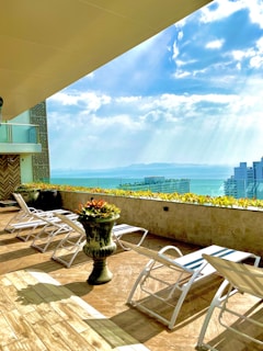 A rooftop terrace features multiple white lounge chairs spread on a wooden floor. A large decorative planter with vibrant flowers is centrally placed. The terrace railing overlooks a scenic view of the sea and distant land. The sky is bright with scattered clouds and sunlight streaming through them.