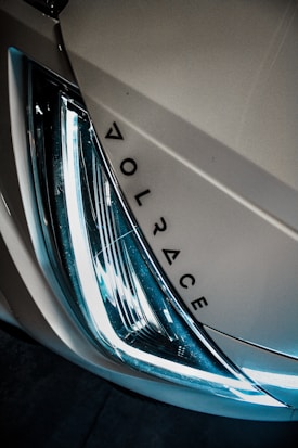 A close-up view of a vehicle's headlight with sleek, modern design elements. The headlight is illuminated with a bright, white light, and there is a stylized logo or brand name next to it, reading 'VOLRACE'. The image has a moody and dramatic lighting effect.