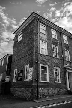A tall brick building with multiple windows appears to be a historical or older structure. It has a street sign indicating a school zone with a speed limit. The sky is partly cloudy, creating a dramatic atmosphere in the black-and-white imagery.