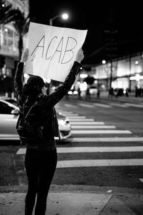 A person stands on a city street holding a large sign above their head with bold letters reading 'ACAB'. The scene is captured at night with blurred streetlights and buildings in the background, suggesting a busy urban environment. The individual is dressed in dark clothing, including a jacket and backpack.