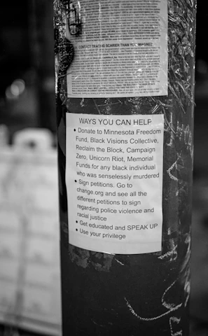 A black and white poster is taped to a pole, containing text about ways to help social justice causes. The poster mentions donating to various funds, signing petitions, getting educated on issues, and using personal privilege to speak up.