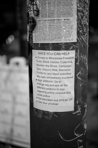 A black and white poster is taped to a pole, containing text about ways to help social justice causes. The poster mentions donating to various funds, signing petitions, getting educated on issues, and using personal privilege to speak up.