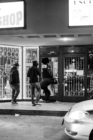 A group of individuals wearing hoodies is gathered outside a storefront at night. One person is kneeling and appears to be interacting with a window or door that is broken, as scattered debris is visible on the ground. The store is closed, barred, and locked, with various signs displayed on the doors and windows. A car is partially visible at the edge of the frame.