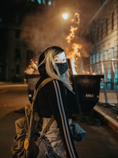 woman in black and white mask and brown jacket standing on sidewalk during night time