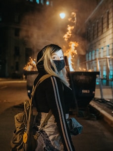 woman in black and white mask and brown jacket standing on sidewalk during night time