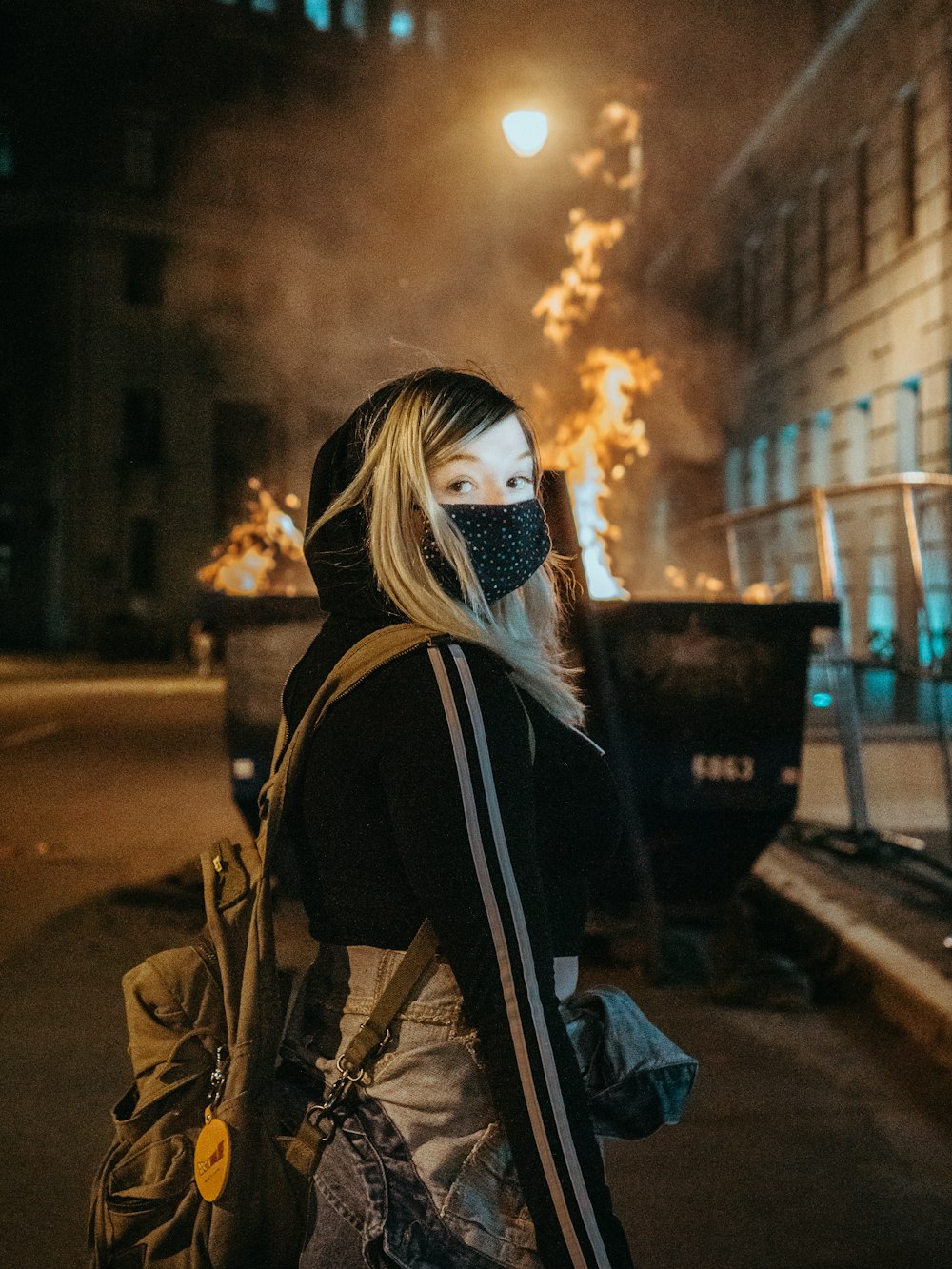 Frau in schwarz-weißer Maske und brauner Jacke steht nachts auf dem Bürgersteig