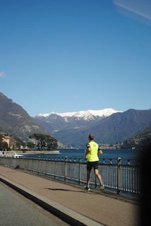 A person wearing a bright yellow shirt is jogging along a waterfront path, with tall, snow-capped mountains and a large body of water in the background. The sky is clear and blue, contributing to a serene atmosphere.