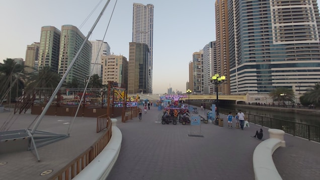 A modern urban waterfront area featuring tall residential and commercial skyscrapers lining a canal. The scene includes a walkway with people strolling, some with strollers, and visible palm trees adding a touch of greenery. Decorative lighting and outdoor dining areas are present, suggesting a leisure and recreational ambiance.