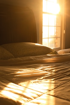 Morning sunlight streams through a window, casting golden light across a neatly made bed with white sheets and a floral-patterned pillow. The room feels warm and serene, with the sunlight casting distinct shadows and highlights on the bed.