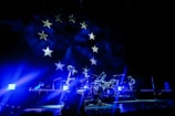 A band performs on stage under vibrant blue lighting, creating a dramatic atmosphere. The backdrop features stars, reminiscent of a flag design, adding to the visual impact of the scene. The musicians appear silhouetted due to the intense lighting coming from behind.