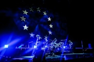 A band performs on stage under vibrant blue lighting, creating a dramatic atmosphere. The backdrop features stars, reminiscent of a flag design, adding to the visual impact of the scene. The musicians appear silhouetted due to the intense lighting coming from behind.