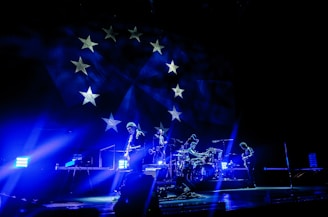 A band performs on stage under vibrant blue lighting, creating a dramatic atmosphere. The backdrop features stars, reminiscent of a flag design, adding to the visual impact of the scene. The musicians appear silhouetted due to the intense lighting coming from behind.
