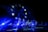 A band performs on stage under vibrant blue lighting, creating a dramatic atmosphere. The backdrop features stars, reminiscent of a flag design, adding to the visual impact of the scene. The musicians appear silhouetted due to the intense lighting coming from behind.