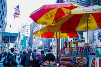 Tourists and locals gather around a hot dog stand in a bustling city street. Bright red and yellow umbrellas display branding and advertisements for beef sausages and hot dogs. A variety of drinks and food items are visible in the stand. Skyscrapers and an American flag are seen in the background, indicating a central urban location.