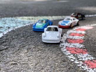 Four toy cars are positioned on a textured asphalt surface, resembling a racetrack. The cars are brightly colored in shades of white, blue, green, and red. They are arranged beside painted lines that mimic race track markings in white and red.