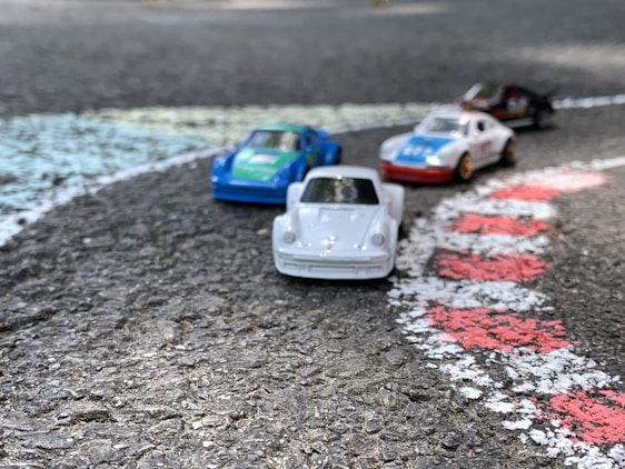 Four toy cars are positioned on a textured asphalt surface, resembling a racetrack. The cars are brightly colored in shades of white, blue, green, and red. They are arranged beside painted lines that mimic race track markings in white and red.