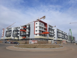 A modern apartment complex under construction with multiple cranes visible. The building features a mix of white, grey, and bright orange accents. There are several stories with balconies, and the area is surrounded by a construction site with barriers and signs. A few cars are parked on the street nearby, and there is a circular road with directional signs at the intersection.