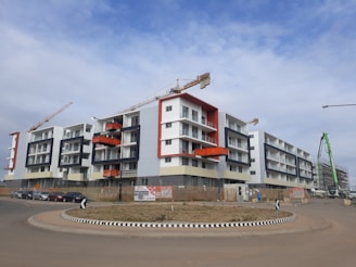 A modern apartment complex under construction with multiple cranes visible. The building features a mix of white, grey, and bright orange accents. There are several stories with balconies, and the area is surrounded by a construction site with barriers and signs. A few cars are parked on the street nearby, and there is a circular road with directional signs at the intersection.