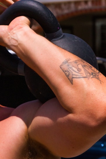 A muscular arm with a tattoo is lifting a black kettlebell, highlighting strength and physical activity.