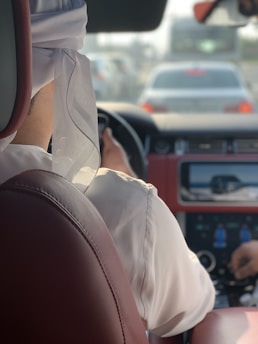 A person wearing a white garment and headscarf is sitting in the driver's seat of a car. The interior features red leather seats, and the view includes the front dashboard and steering wheel. Outside, other vehicles can be seen through the windshield, suggesting the car is in traffic.