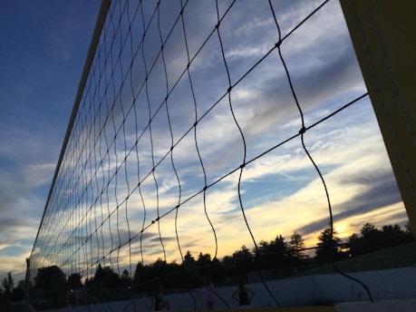A volleyball net stretches across the foreground against a backdrop of a vividly painted sky at sunset. The clouds display a range of colors from blue to orange, with silhouettes of trees and an athletic field visible below.