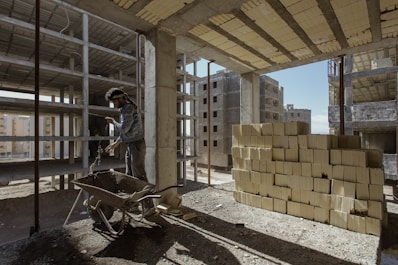 A construction site with multiple unfinished buildings and a worker handling a wheelbarrow. Stacks of building materials, likely concrete blocks, are placed to the side. The area is dusty and lit by natural daylight with shadows cast across the ground.