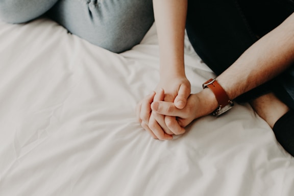 Two individuals are sitting on a white bed, holding hands. One person is wearing a watch with a brown leather strap, and both are casually dressed in comfortable clothing.