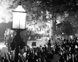 A black and white image depicting a protest scene at night. A crowd of people is seen holding up signs and raising their hands, facing a group of police officers in riot gear. A prominent lamppost stands in the foreground, with trees surrounding the area. The atmosphere is tense, with smoke or tear gas visible in the air.