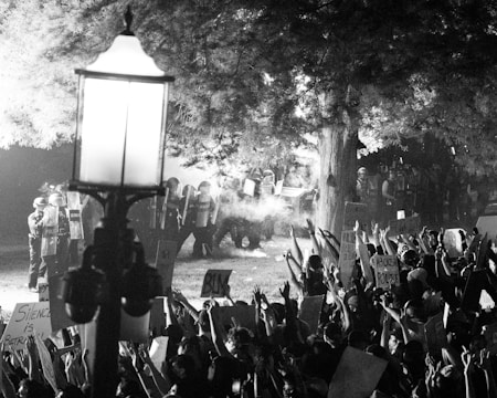 A black and white image depicting a protest scene at night. A crowd of people is seen holding up signs and raising their hands, facing a group of police officers in riot gear. A prominent lamppost stands in the foreground, with trees surrounding the area. The atmosphere is tense, with smoke or tear gas visible in the air.