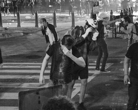 A group of people, some of whom appear to be in distress, gather on a street with a zebra crossing. One person pours water on their face while others wipe their faces. There is visible chaos in the background, with more people and scattered debris.