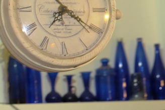A vintage clock with Roman numerals is prominently displayed in the foreground. The background features a collection of blue glass bottles and vases arranged on a shelf.