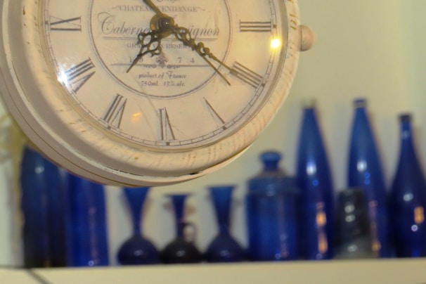A vintage clock with Roman numerals is prominently displayed in the foreground. The background features a collection of blue glass bottles and vases arranged on a shelf.