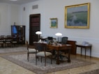 A well-furnished office space with a large wooden desk featuring several items including lamps and a computer. The walls are adorned with framed paintings. There is a meeting area with chairs on the left side and a wooden bookshelf integrated into the wall. The room has a classic and elegant design with patterned rugs on the floor.