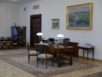 A well-furnished office space with a large wooden desk featuring several items including lamps and a computer. The walls are adorned with framed paintings. There is a meeting area with chairs on the left side and a wooden bookshelf integrated into the wall. The room has a classic and elegant design with patterned rugs on the floor.