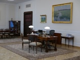 A well-furnished office space with a large wooden desk featuring several items including lamps and a computer. The walls are adorned with framed paintings. There is a meeting area with chairs on the left side and a wooden bookshelf integrated into the wall. The room has a classic and elegant design with patterned rugs on the floor.