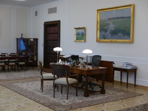 A well-furnished office space with a large wooden desk featuring several items including lamps and a computer. The walls are adorned with framed paintings. There is a meeting area with chairs on the left side and a wooden bookshelf integrated into the wall. The room has a classic and elegant design with patterned rugs on the floor.