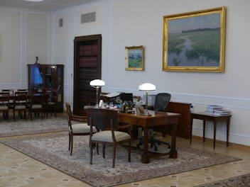 A well-furnished office space with a large wooden desk featuring several items including lamps and a computer. The walls are adorned with framed paintings. There is a meeting area with chairs on the left side and a wooden bookshelf integrated into the wall. The room has a classic and elegant design with patterned rugs on the floor.