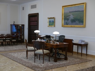 A well-furnished office space with a large wooden desk featuring several items including lamps and a computer. The walls are adorned with framed paintings. There is a meeting area with chairs on the left side and a wooden bookshelf integrated into the wall. The room has a classic and elegant design with patterned rugs on the floor.