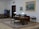 A well-furnished office space with a large wooden desk featuring several items including lamps and a computer. The walls are adorned with framed paintings. There is a meeting area with chairs on the left side and a wooden bookshelf integrated into the wall. The room has a classic and elegant design with patterned rugs on the floor.