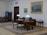 A well-furnished office space with a large wooden desk featuring several items including lamps and a computer. The walls are adorned with framed paintings. There is a meeting area with chairs on the left side and a wooden bookshelf integrated into the wall. The room has a classic and elegant design with patterned rugs on the floor.