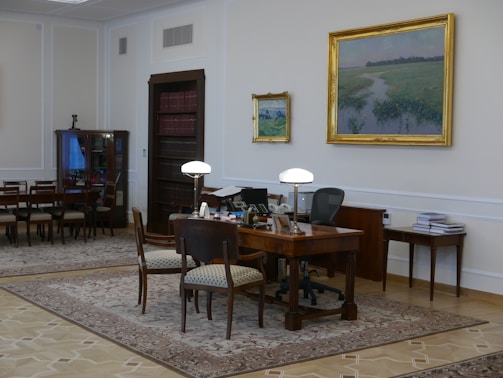 A well-furnished office space with a large wooden desk featuring several items including lamps and a computer. The walls are adorned with framed paintings. There is a meeting area with chairs on the left side and a wooden bookshelf integrated into the wall. The room has a classic and elegant design with patterned rugs on the floor.
