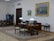 A well-furnished office space with a large wooden desk featuring several items including lamps and a computer. The walls are adorned with framed paintings. There is a meeting area with chairs on the left side and a wooden bookshelf integrated into the wall. The room has a classic and elegant design with patterned rugs on the floor.