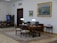 A well-furnished office space with a large wooden desk featuring several items including lamps and a computer. The walls are adorned with framed paintings. There is a meeting area with chairs on the left side and a wooden bookshelf integrated into the wall. The room has a classic and elegant design with patterned rugs on the floor.