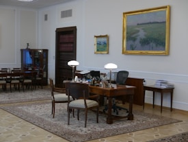 A well-furnished office space with a large wooden desk featuring several items including lamps and a computer. The walls are adorned with framed paintings. There is a meeting area with chairs on the left side and a wooden bookshelf integrated into the wall. The room has a classic and elegant design with patterned rugs on the floor.