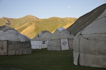 Several yurts are situated on a grassy plain against the backdrop of a mountainous landscape. The scene is bathed in soft evening light, with a clear sky and a moon visible above the horizon.