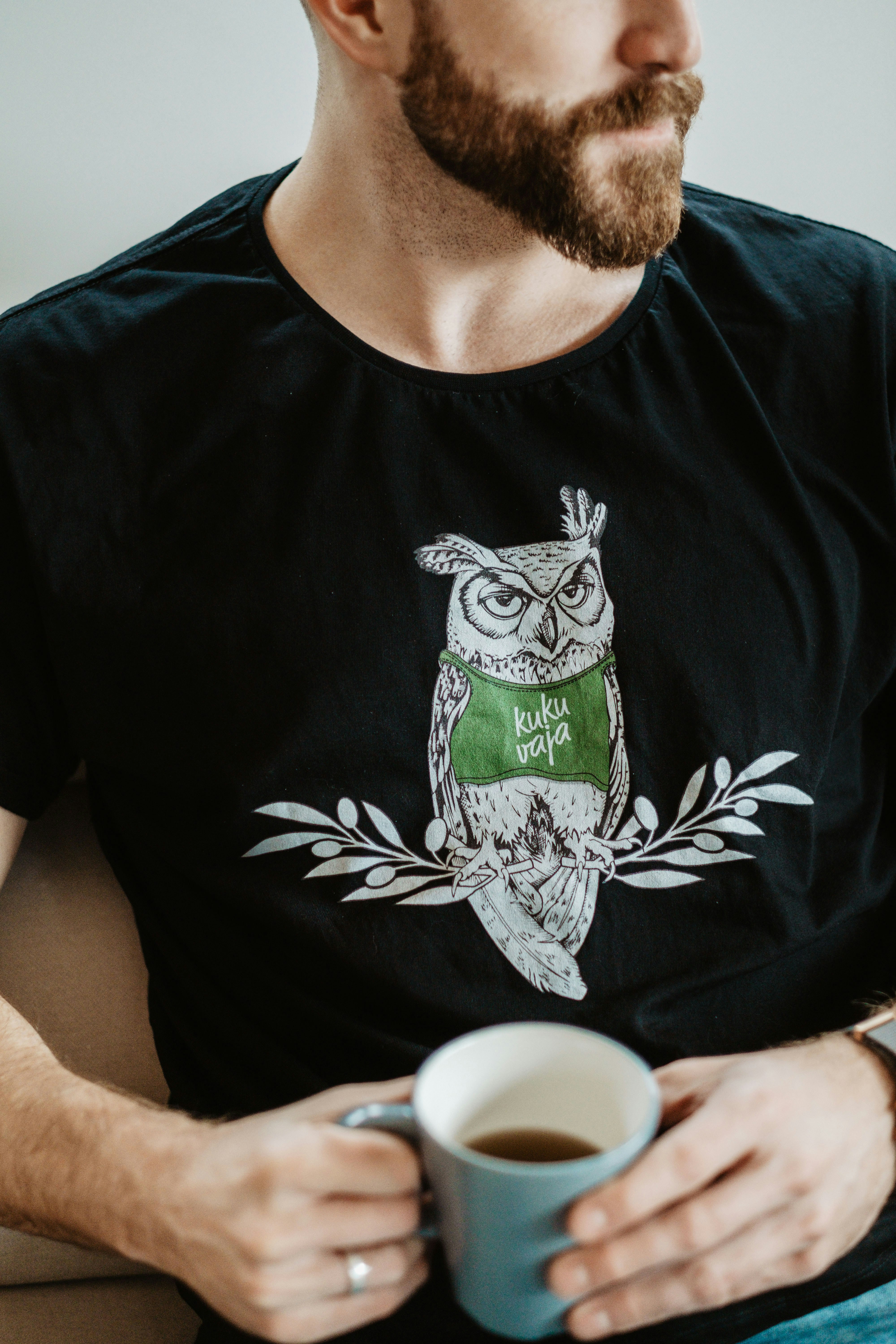 Kukuvaja Merch Owl Logo on Black Shirt with a Cup of Mountaintea