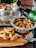 A kitchen scene with freshly cut potato wedges on a wooden cutting board, next to a knife. A stainless steel bowl contains more of the cut wedges, while someone pours oil from a green-labeled bottle over them. A wire basket and a decorative wooden item are in the background along with a container holding tomatoes.
