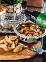 A kitchen scene with freshly cut potato wedges on a wooden cutting board, next to a knife. A stainless steel bowl contains more of the cut wedges, while someone pours oil from a green-labeled bottle over them. A wire basket and a decorative wooden item are in the background along with a container holding tomatoes.