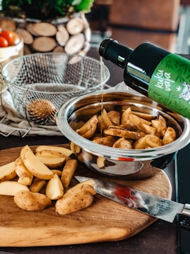A kitchen scene with freshly cut potato wedges on a wooden cutting board, next to a knife. A stainless steel bowl contains more of the cut wedges, while someone pours oil from a green-labeled bottle over them. A wire basket and a decorative wooden item are in the background along with a container holding tomatoes.