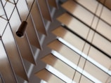 A close-up view of horizontal wooden blinds with light coming through the gaps, creating geometric patterns. A dark brown cord with a wooden pull hangs in the foreground, adding depth to the composition.